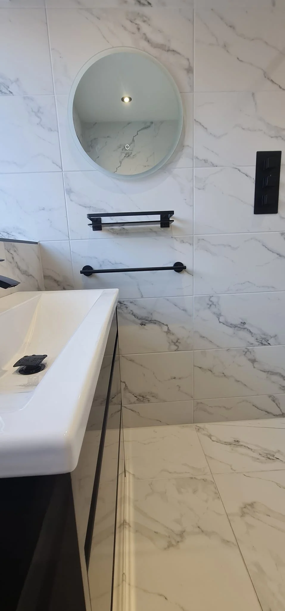 Modern bathroom with white marble walls and floor, black towel racks, a small round mirror, a white rectangular sink with black legs, and a black triple switch on the wall.