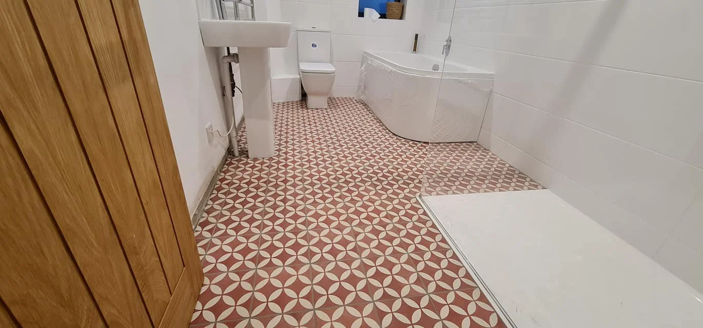 Bathroom with pink and white geometric patterned tile floor, a white toilet, small sink, white bathtub, and a wooden door.