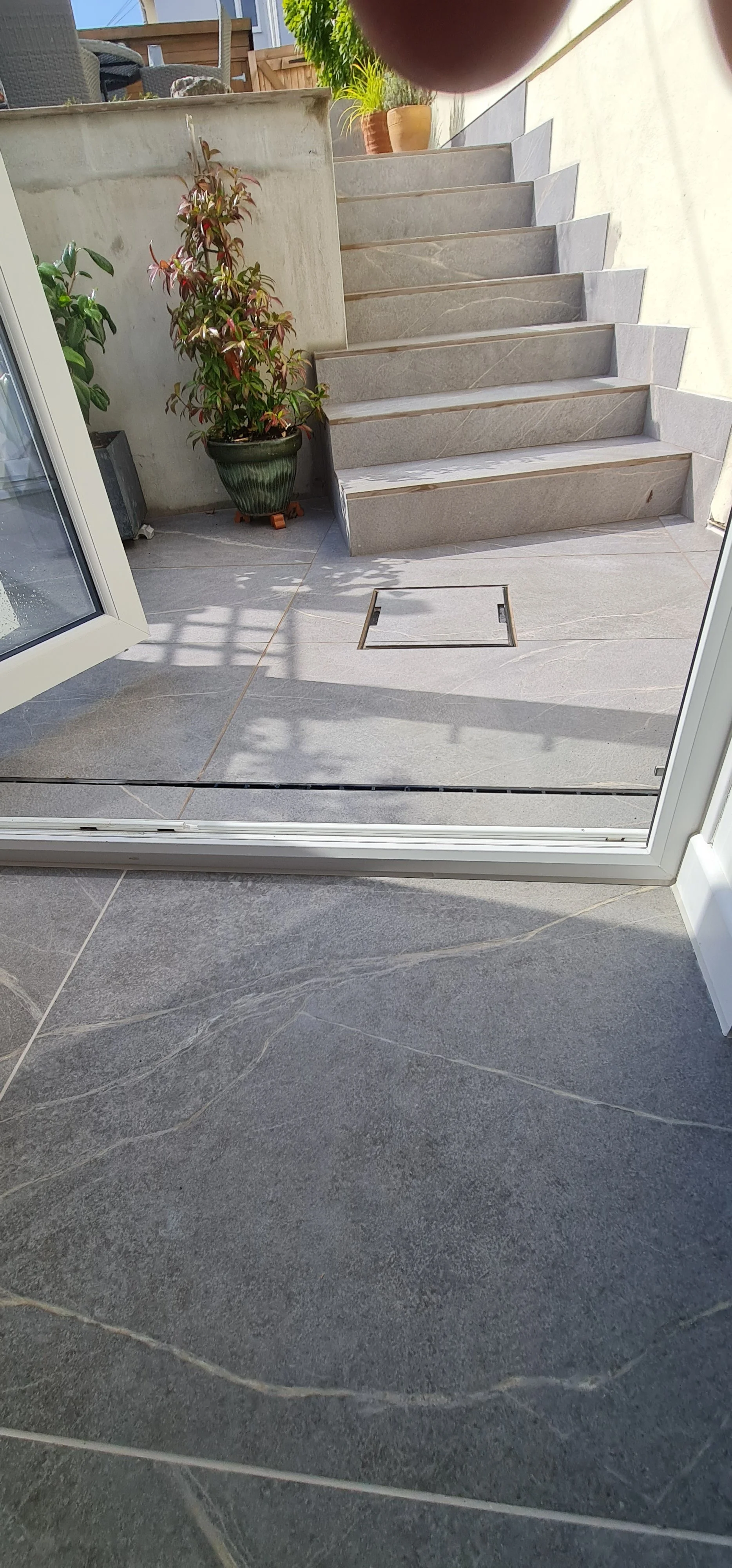 View of outdoor patio with tiled floor, potted plants, and stone staircase leading to an upper level, visible from inside through a glass door.