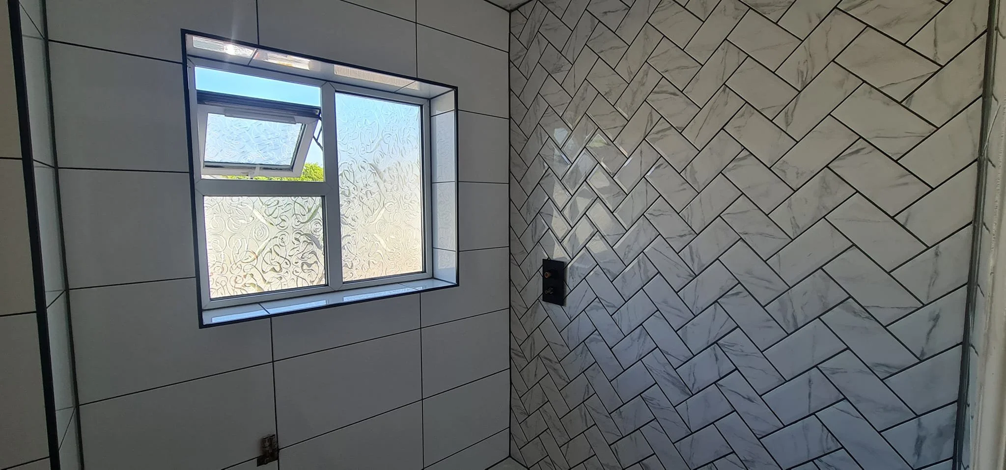 A room under construction or renovation with tiled walls and a window, showing a partly opened window with patterned frosted glass, and black fixtures on the wall.