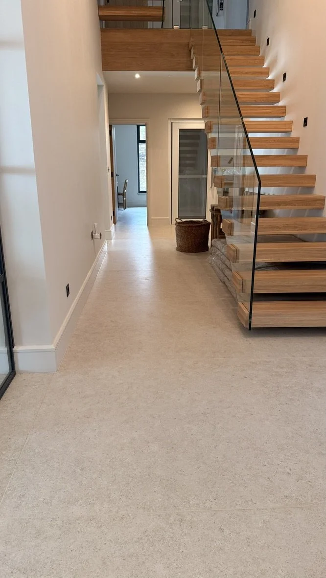 Interior view of a modern home with a staircase featuring wooden steps and glass railing, beige flooring, white walls, and a basket near the staircase.
