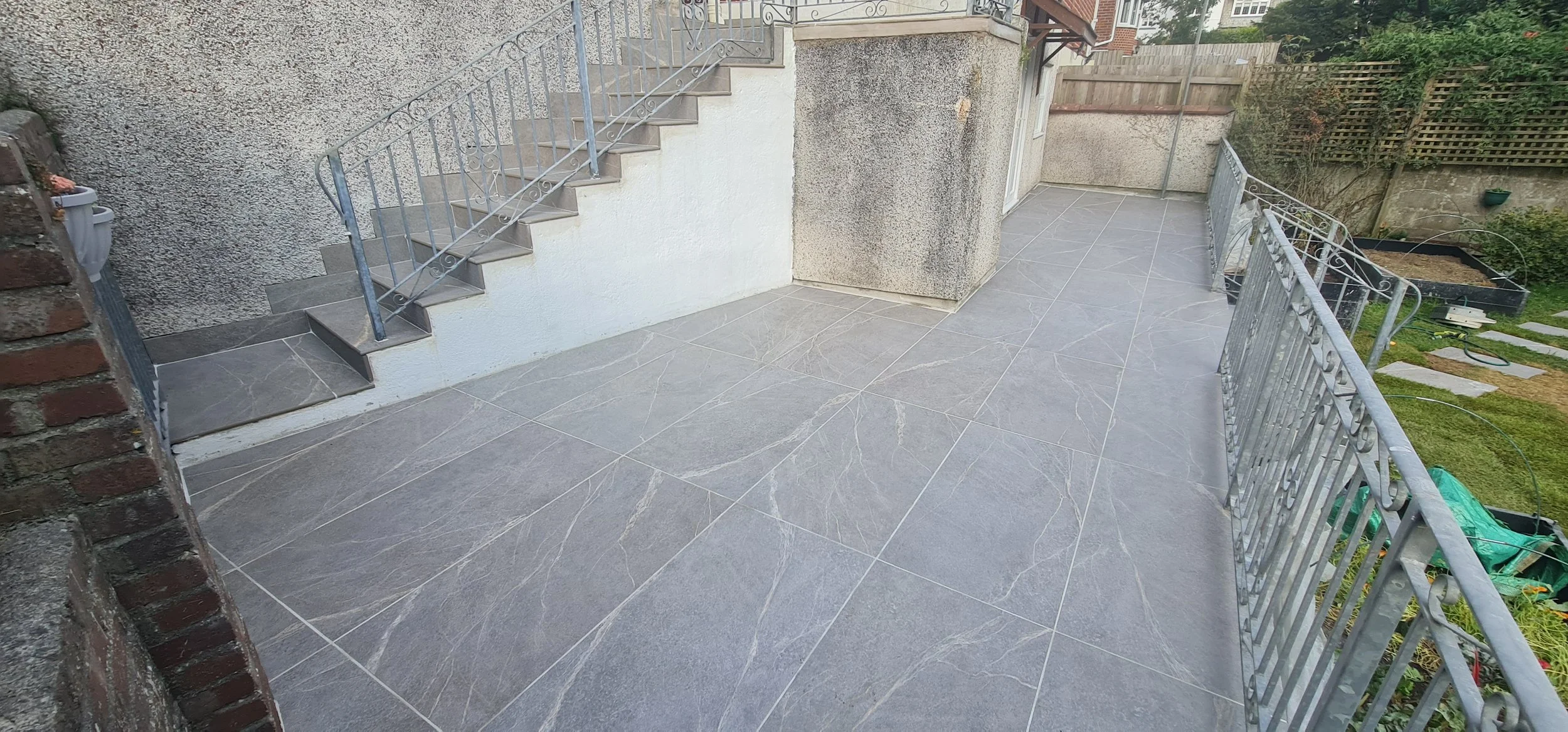 Empty outdoor patio with gray tiled flooring, metal railing, stairs leading to an upper level, and a garden area with green grass and plants in the background.