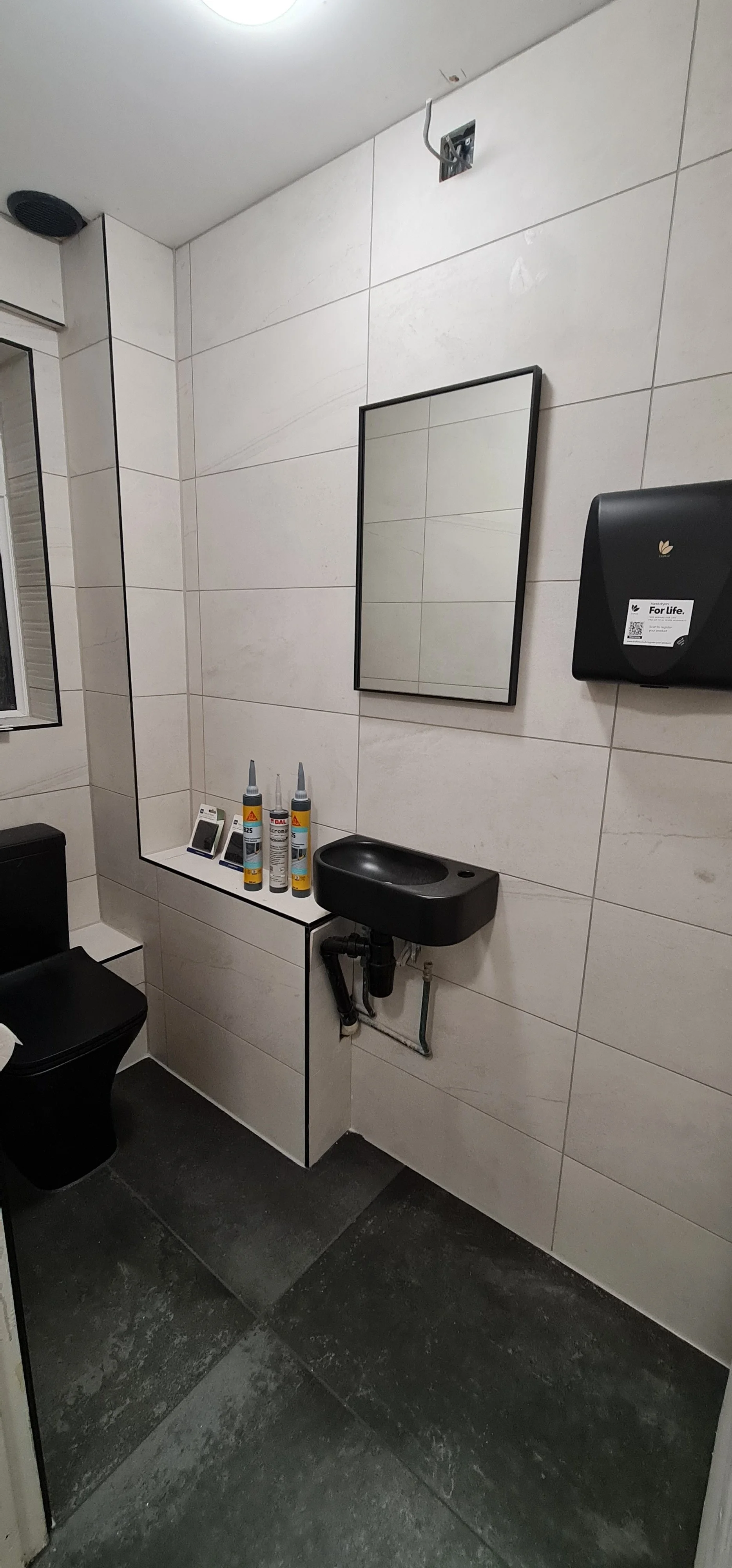 A small bathroom with a black toilet, a black sink, a mirror, a black wall-mounted paper towel dispenser, and some bottles of adhesive glue on a white ledge. There is a small window and a vent in the ceiling.
