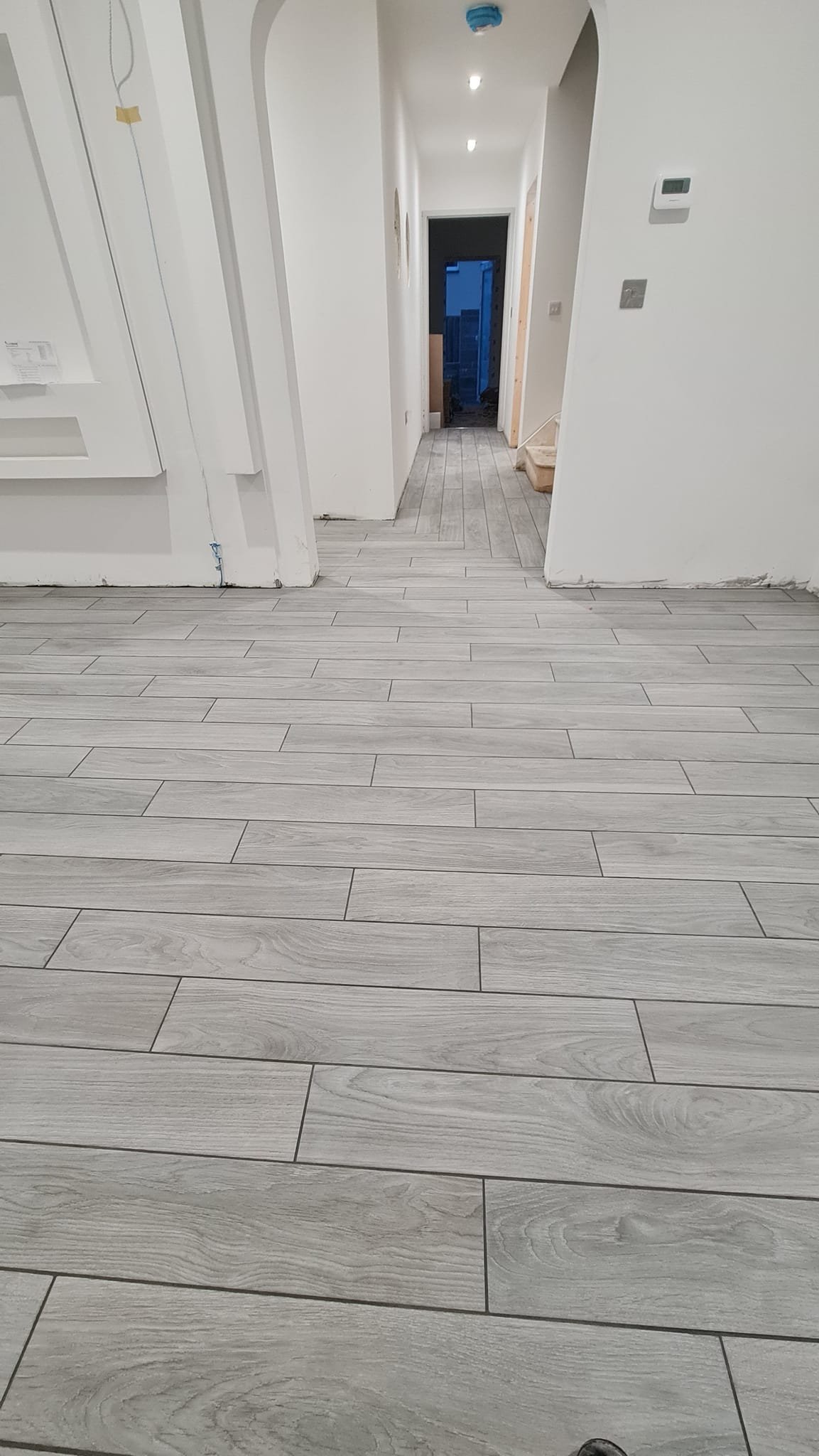 Interior hallway with light-colored wooden floor tiles, white walls, and a doorway at the end leading outside. Some construction or renovation materials are visible near the doorway.