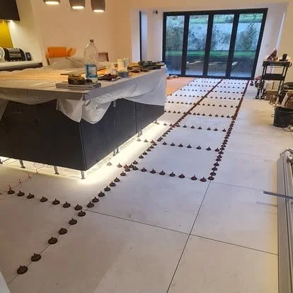 A room with a large table covered with various items, and chocolate candies arranged in a grid pattern on the floor, leading toward a sliding glass door showing an outdoor view.