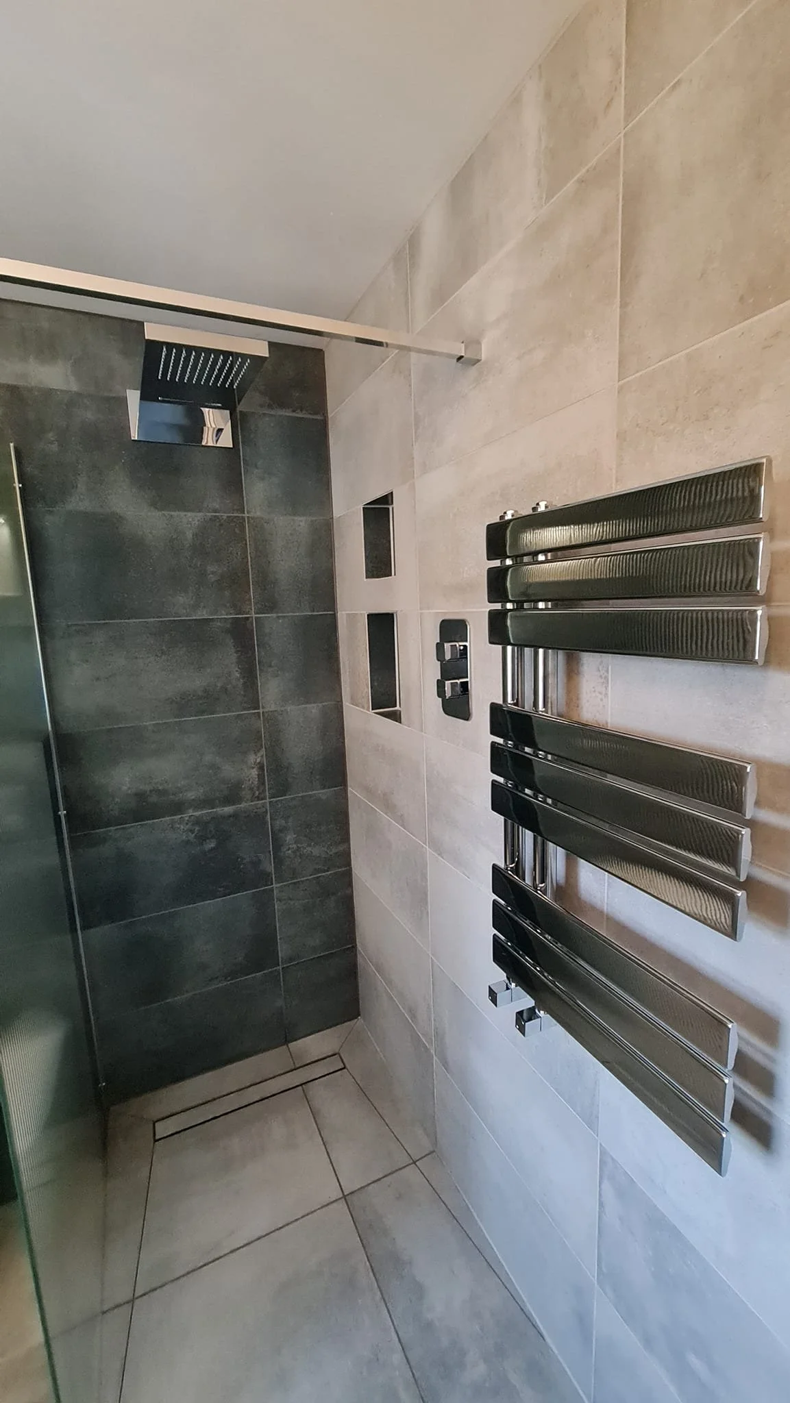 Interior view of a modern bathroom, featuring a heated towel rail, a tiled wall and floor, a shower area with dark tiles, and built-in wall niches.