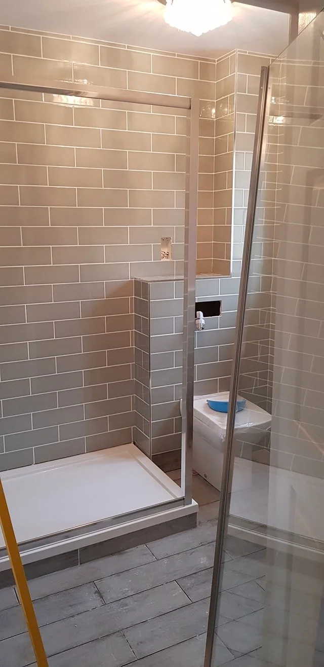 A bathroom under construction with gray tiled walls, a small shower area with a white base, and a toilet covered with a plastic sheet. There are metal fixtures and a partially open glass shower door.