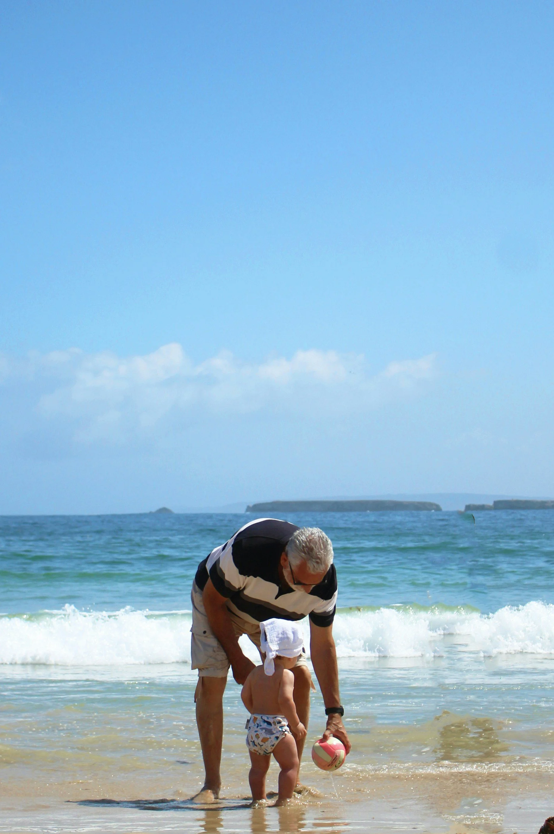 An older man and a young child at the beach, playing with a beach ball near the shoreline, with ocean and distant cliffs visible in the background on a sunny day.