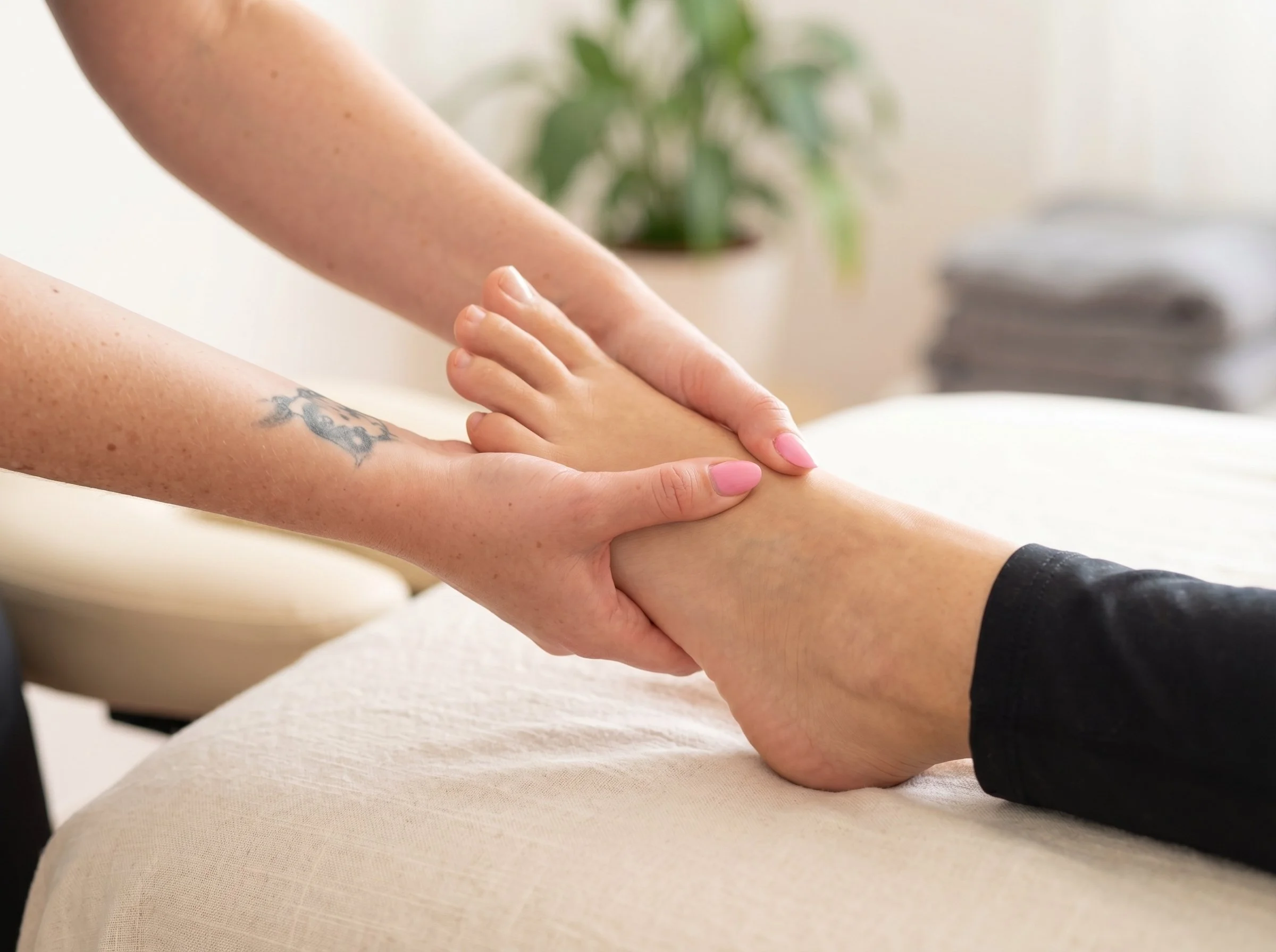 Close-up of a person receiving a massage, showing their arm and hand being gently massaged with a second person's hands, with a blurred background including a plant.