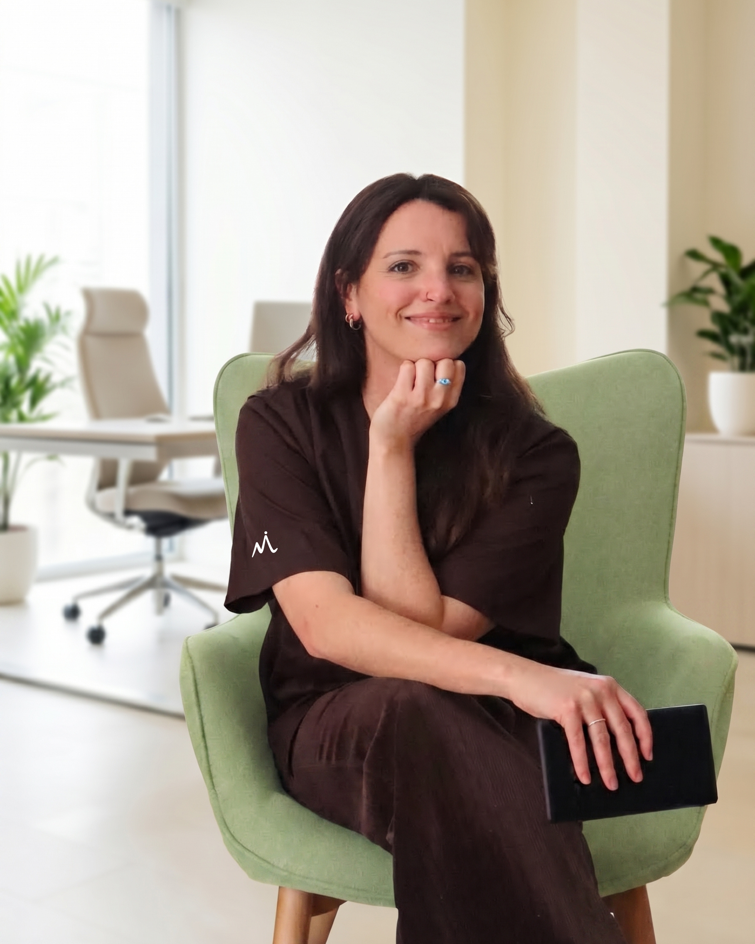 A woman with dark hair smiling, sitting in a green chair in a bright office setting with white walls, a desk, and potted plants in the background.