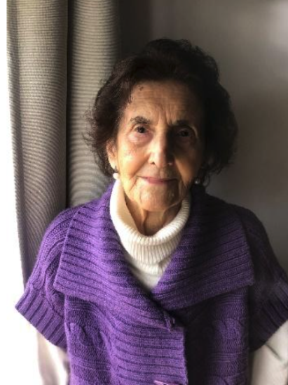 An elderly woman with dark curly hair, dressed in a white turtleneck and purple sweater, sitting near a window with curtains.