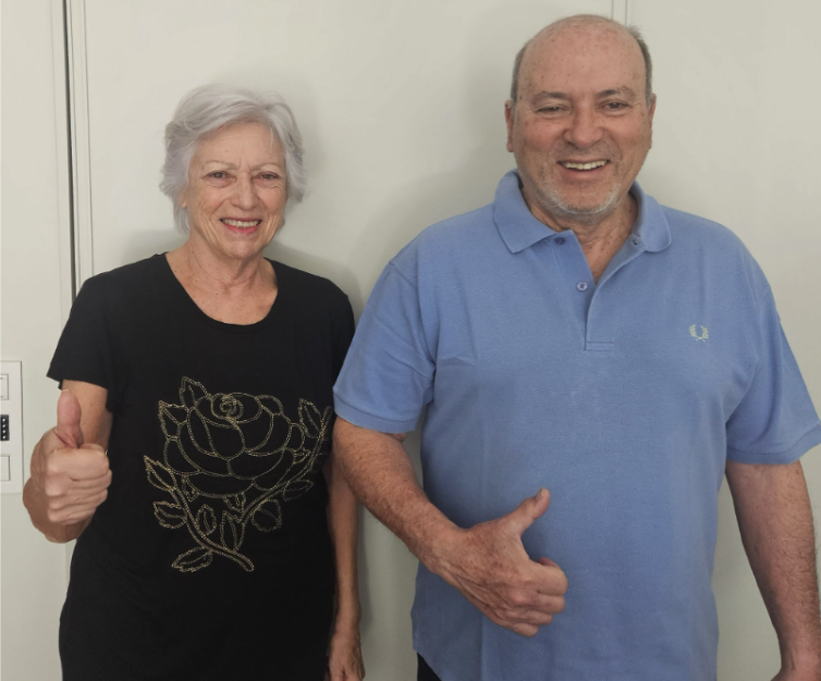 An elderly woman with short gray hair wearing a black t-shirt with a gold rose design and an elderly man with a shaved head wearing a light blue polo shirt, both smiling and giving thumbs up in front of a plain wall.