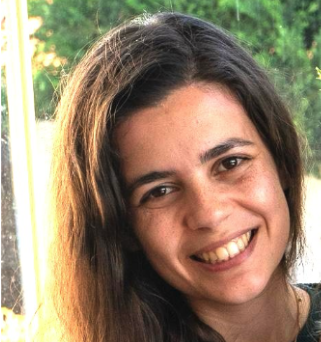 Close-up of a young woman with long dark hair, smiling outdoors with greenery in the background.