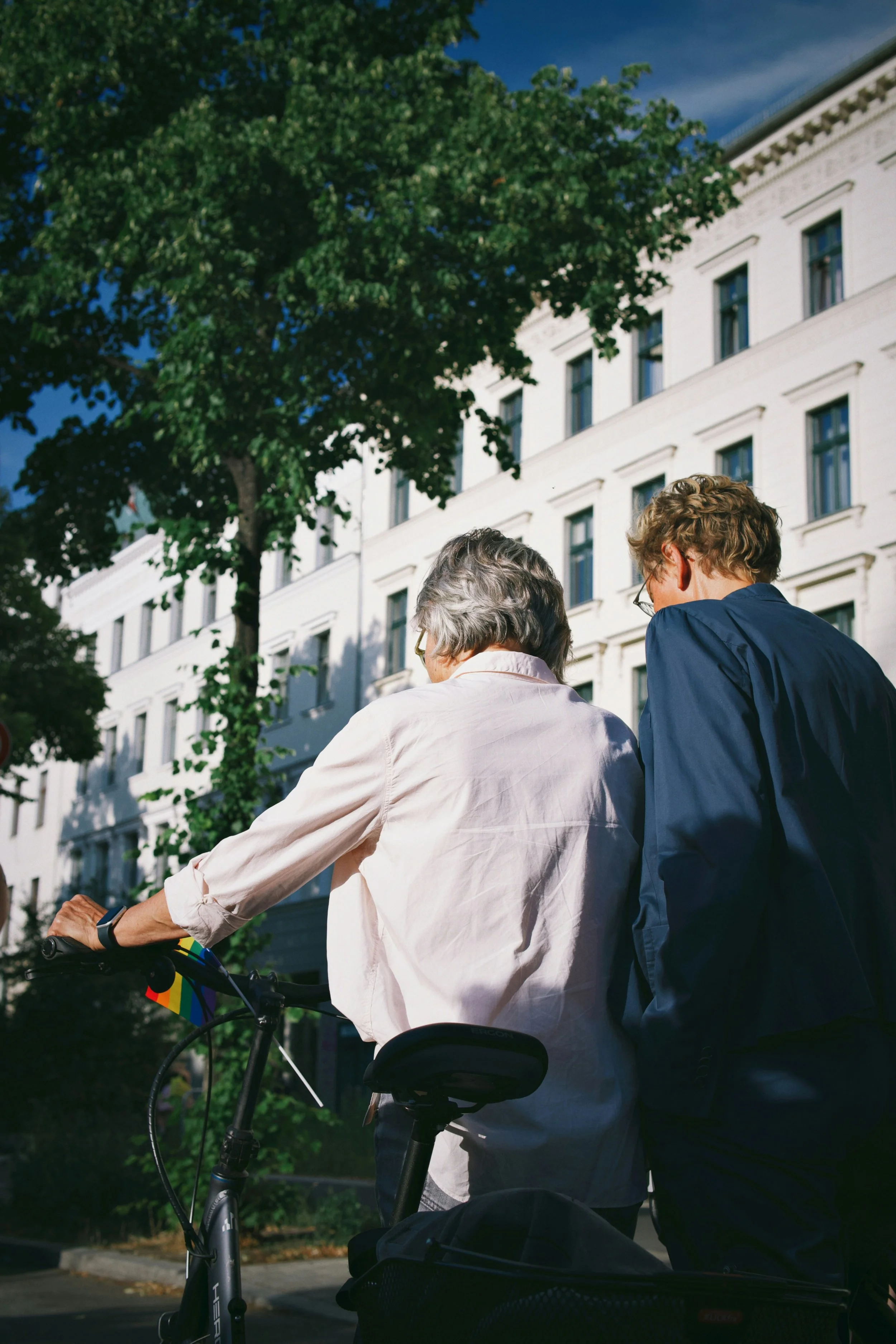 Two people walking with bicycles in an urban area, with a large white building and green trees in the background.