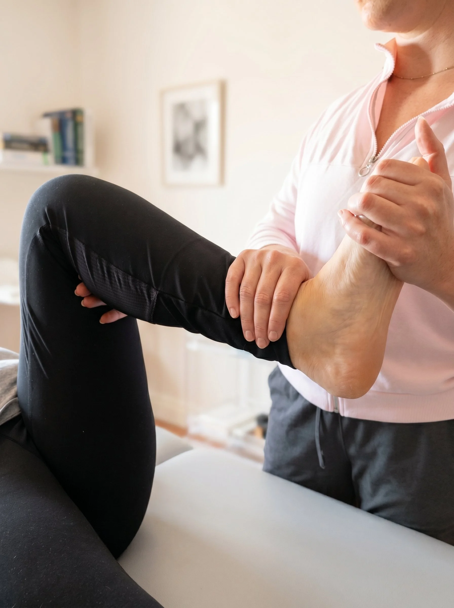 A person is performing a physical therapy exercise, holding another person's hand with one hand and supporting their wrist with the other, while sitting on a white surface in a room, with a blurred shelf and picture in the background.