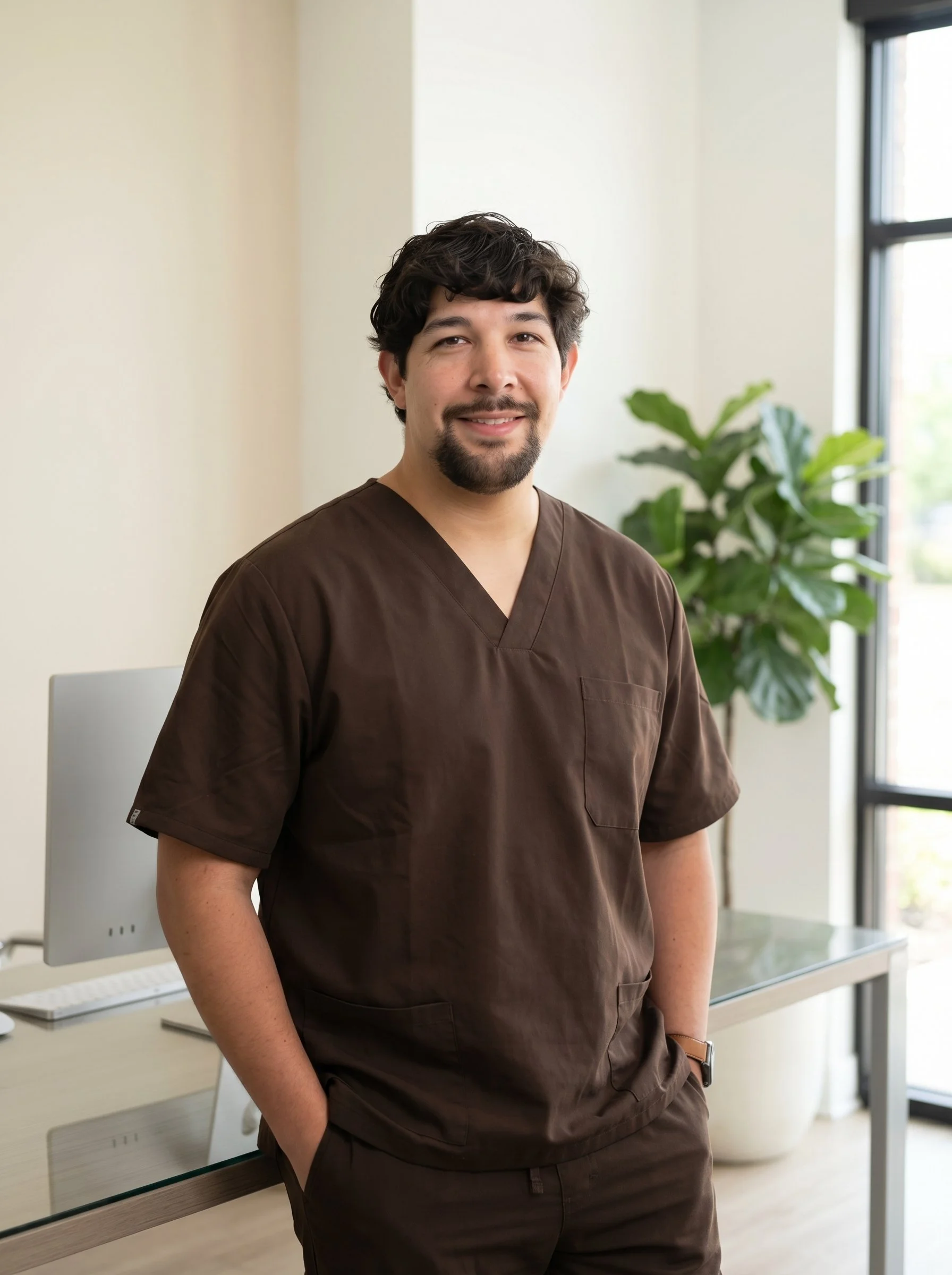 A man in a brown medical uniform sitting in a modern, well-lit office space with large windows and green plants.