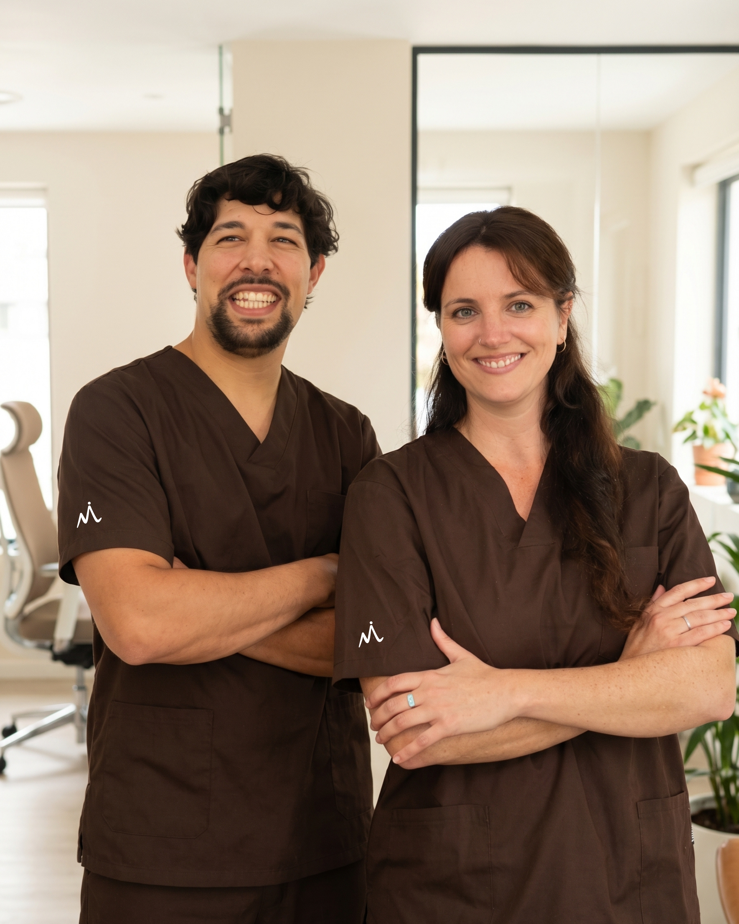 Two medical professionals, a man and a woman, standing side by side in a medical office or clinic, smiling and looking at the camera, with a mirror and office furniture visible in the background.