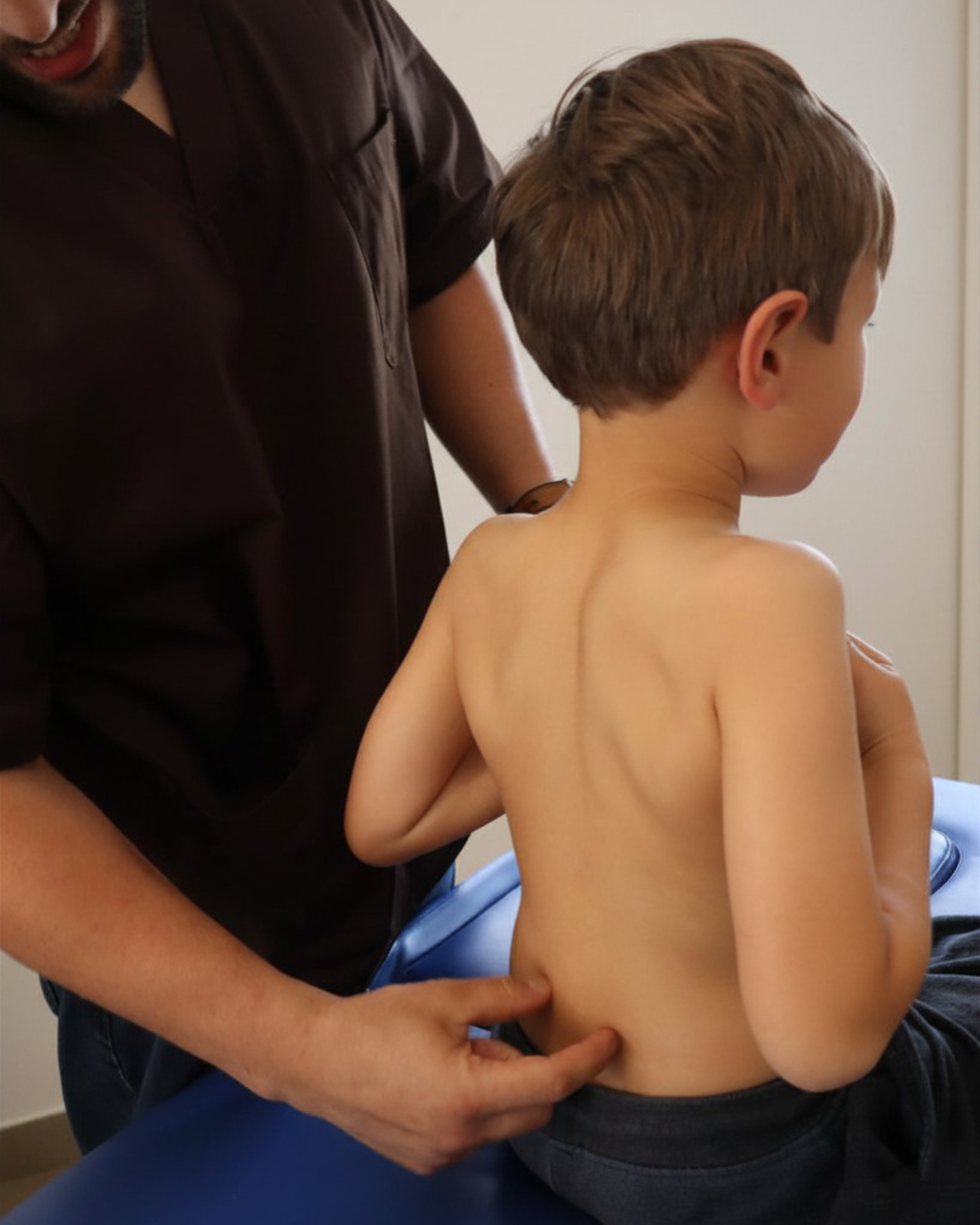A person with a beard wearing a dark shirt is standing behind a shirtless young boy with light brown hair. The person is holding the boy's waist with one hand, gently supporting him.