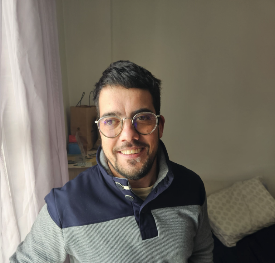 A man with dark hair and glasses smiling indoors near a window with curtains, with a bed and pillow visible in the background.
