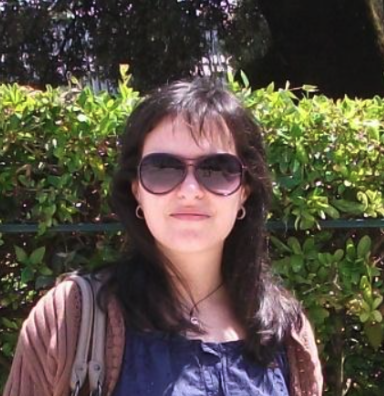 A woman with dark hair, wearing sunglasses, a blue top, and a brown jacket, standing outdoors in front of green bushes.