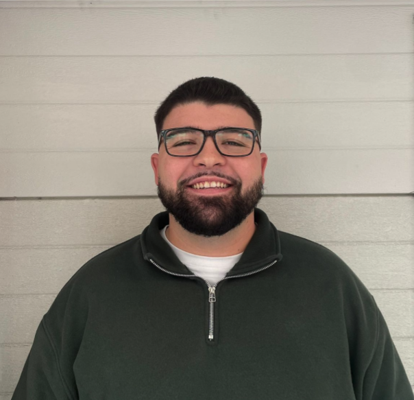 A man with dark hair, glasses, a beard, wearing a dark green zip-up jacket, smiling in front of a white wooden wall.