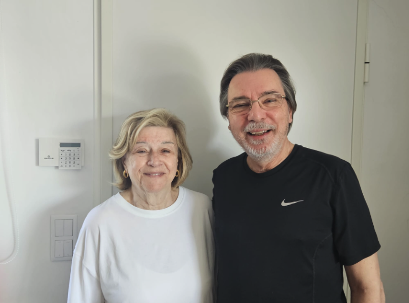 Smiling elderly woman with blonde hair wearing a white shirt, standing next to smiling middle-aged man with dark hair and glasses, wearing a black Nike t-shirt.