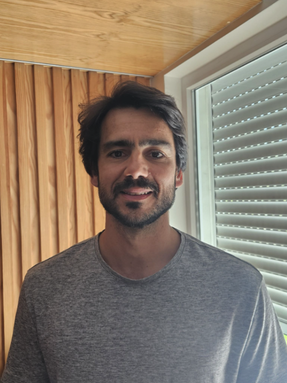 A man with dark hair and a beard smiling indoors, standing in front of a window with closed blinds and wooden paneling on the wall behind him.