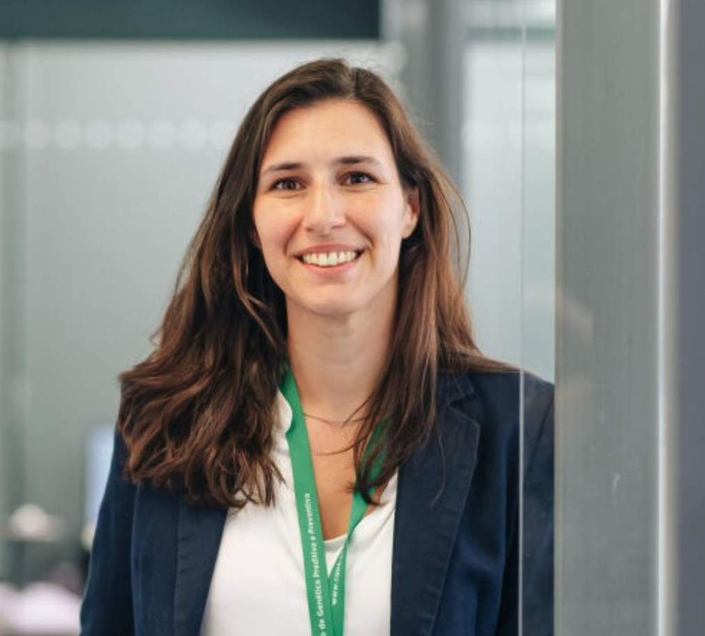 A woman with brown hair smiling, wearing a dark blazer and a green lanyard, standing indoors near glass walls.