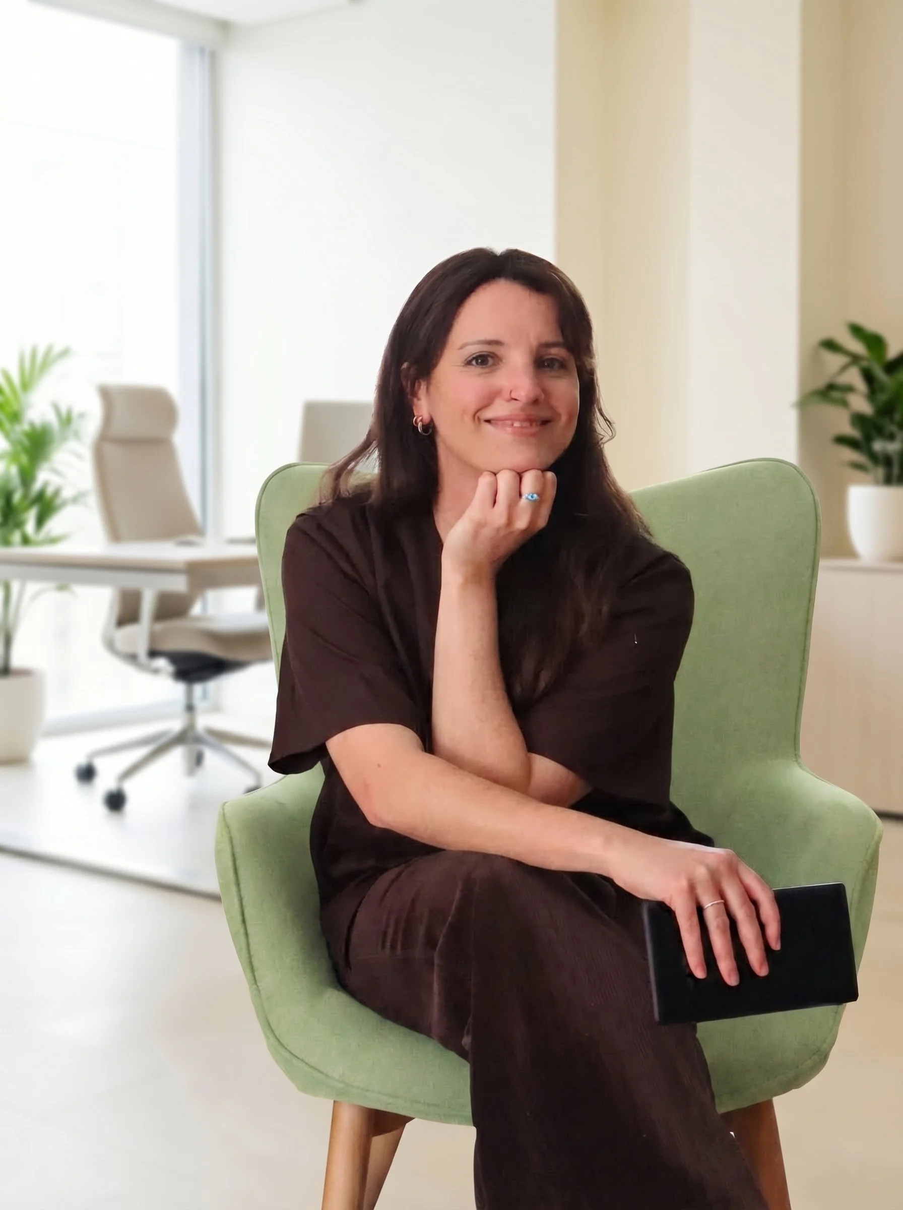 A woman with dark hair, wearing a black top, sitting in a lime green chair in a modern office environment with white walls, glass windows, and plants.