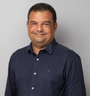 A smiling man with short dark hair, wearing a navy blue button-up shirt, standing against a gray background.