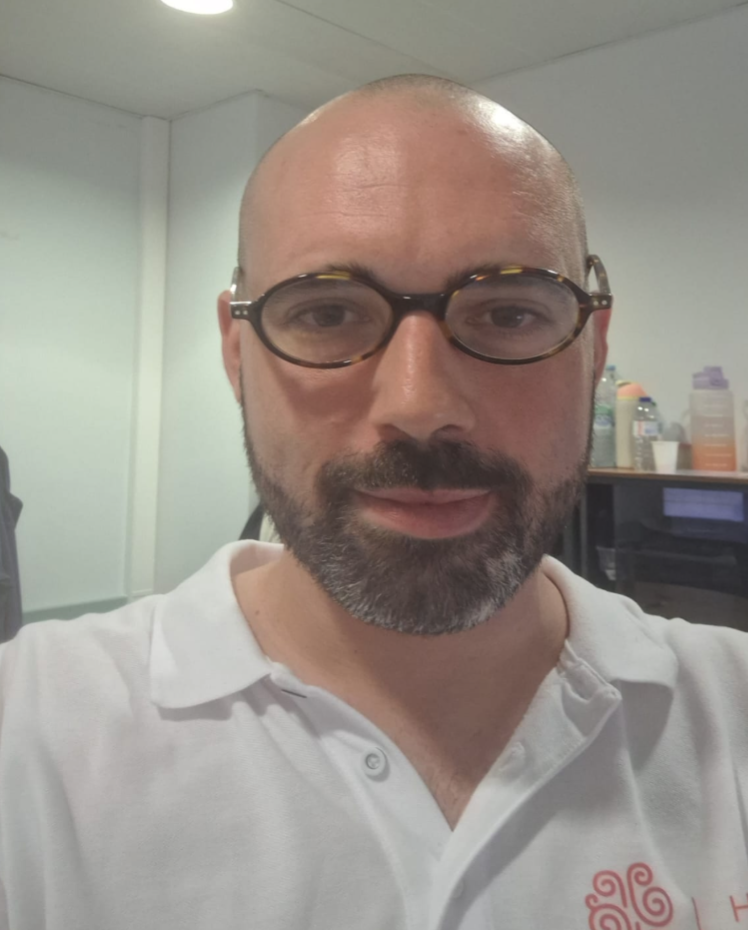 A man with glasses, a bald head, and a beard wearing a white polo shirt taking a selfie in an office or break room with shelves and bottles in the background.