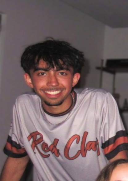 Young man with dark, messy hair and a goatee, smiling, wearing a white T-shirt with red and black stripes and the words 'Red Clan' on it.