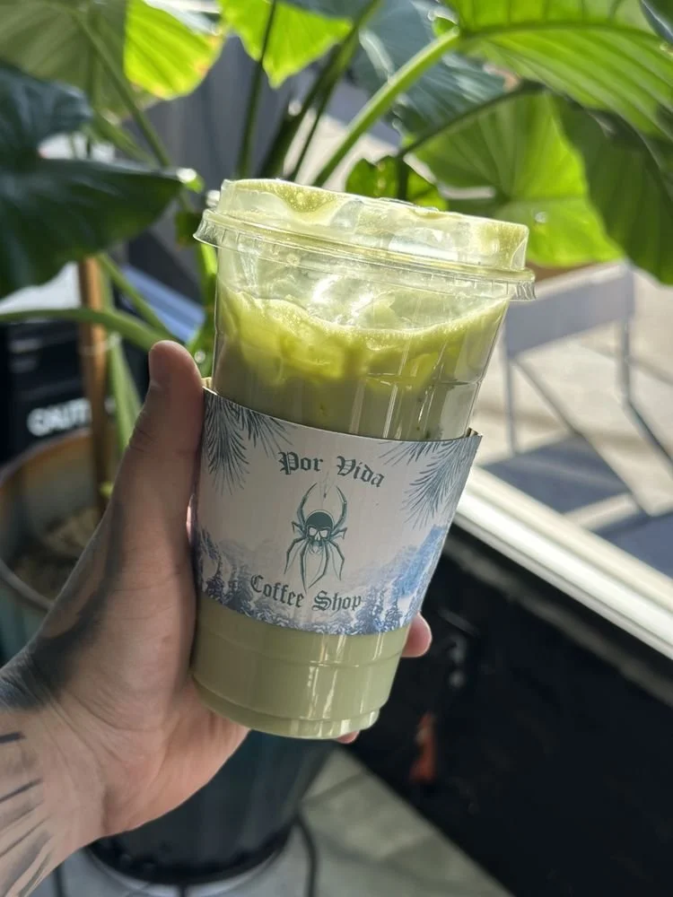 A hand holding a clear plastic cup with a green beverage at Por Vida Coffee Shop, with a label showing a spider logo and tropical leaves, set against a leafy background.