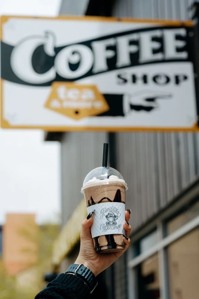 Person holding a chocolate iced coffee or frappuccino with whipped cream and chocolate syrup outside a coffee shop, with a blurred coffee shop sign in the background.