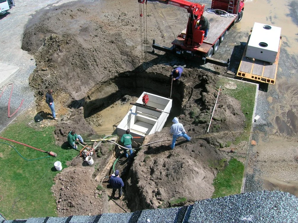 Construction workers installing a large white concrete septic tank in a large open excavation site.