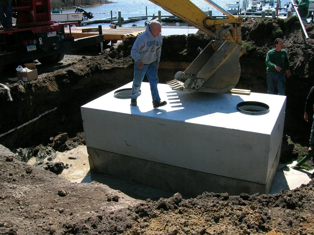 Charlestown,  South Kingstown, Narragansett , Westerly Construction workers installing a large white concrete tank underground near a body of water, with construction equipment and machinery around.