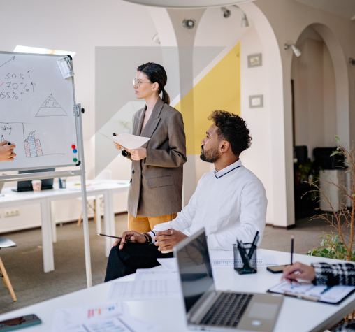 Una mujer de negocios de cabello oscuro y gafas, vestida con traje gris, está de pie junto a una pizarra con gráficos y notas en un entorno de oficina moderna. Un hombre con barba, cabello rizado y camisa blanca está sentado, escuchando atentamente, con una laptop y documentos en la mesa.