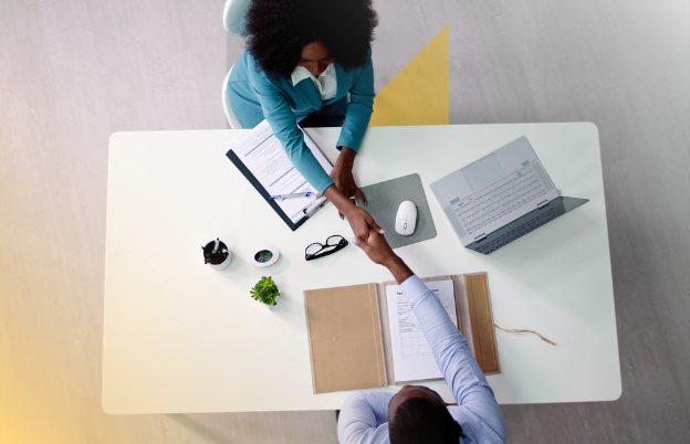 Dos personas en una oficina intercambian un apretón de manos sobre un escritorio, que tiene una computadora portátil, una libreta, y objetos de oficina.