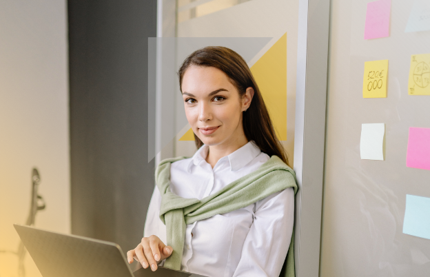 Mujer joven con blusa blanca y suéter verde, sosteniendo una laptop, en un entorno de oficina con notas adhesivas en la pizarra blanca.