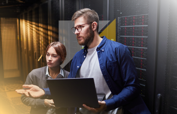 Un hombre y una mujer trabajan en una sala de servidores o data center, con el hombre sosteniendo una laptop y la mujer observando, ambos parecen estar revisando información técnica.