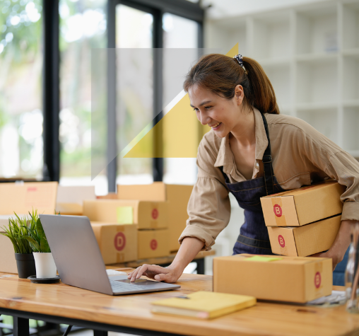 Mujer sonriendo en una oficina de envíos, colocando cajas en un escritorio junto a una laptop.
