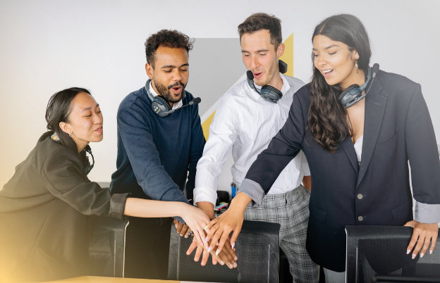Cuatro personas en una mesa de trabajo, con auriculares inalámbricos, colocan sus manos juntas en el centro en señal de unión o colaboración.