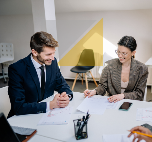 Dos personas en una reunión de trabajo en una oficina, una mujer y un hombre sentados en una mesa con documentos, bolígrafos y un teléfono móvil.