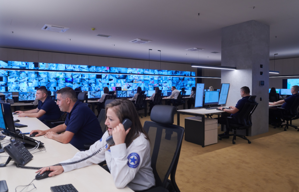 Sala de control con varias personas trabajando en computadoras, en una pared de pantallas de video de fondo, en un ambiente moderno y tecnológico.