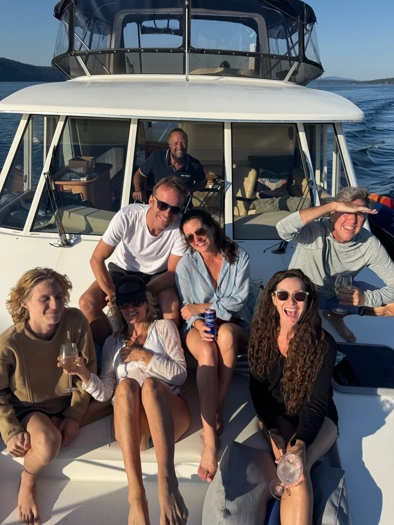 A group of seven people enjoying a day on a boat, sitting and standing on the deck with drinks, with water and distant land in the background.