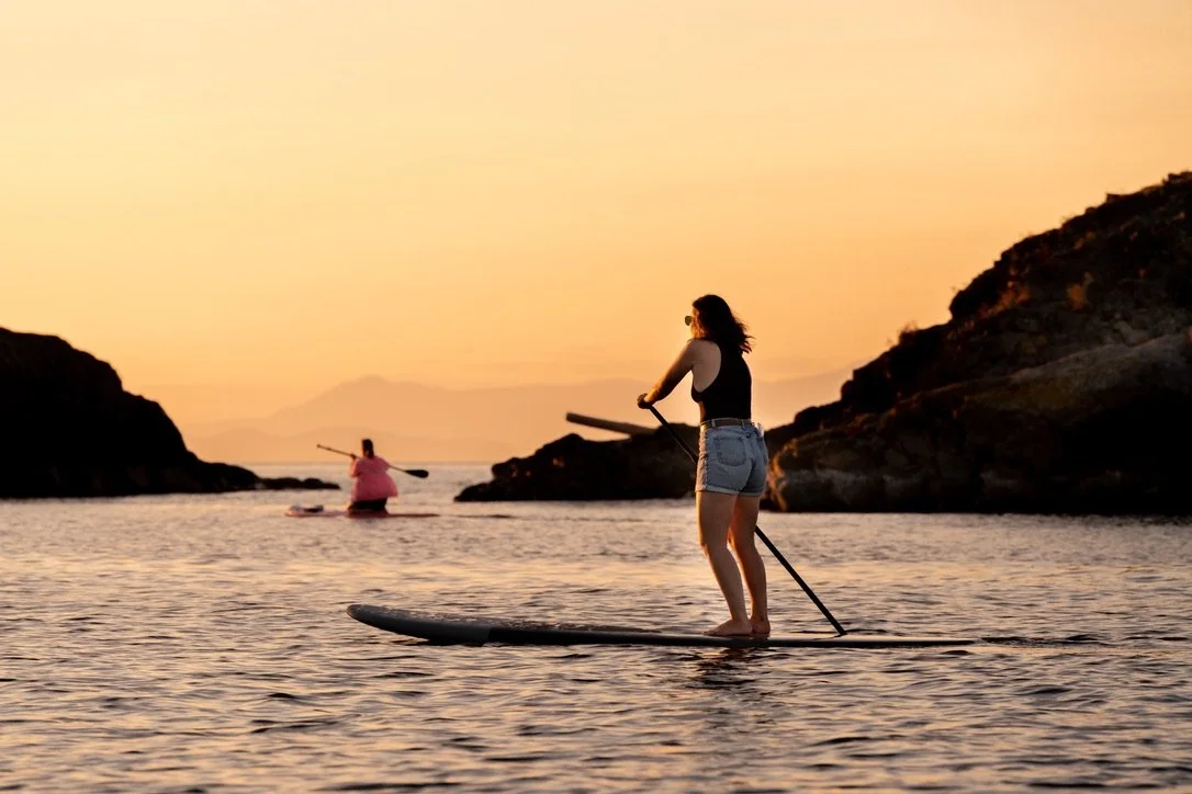 Two women paddleboarding in calm water during sunset with rocky islands in the background.