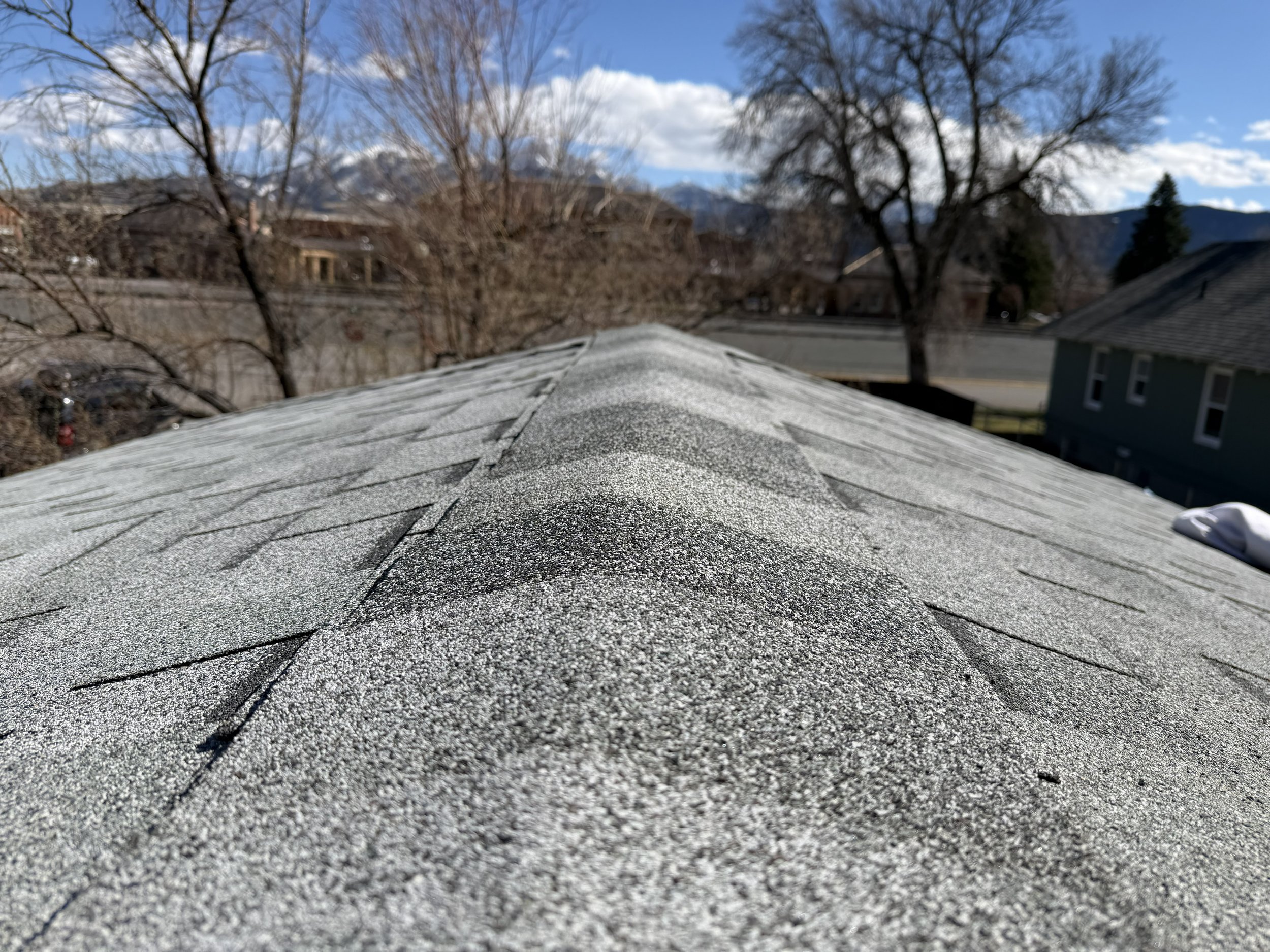 Close-up of a textured gray shingle roof with a background of leafless trees, houses, mountain range, and a partly cloudy sky.