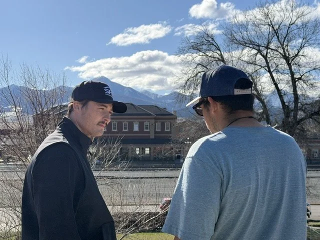 Two men wearing caps and casual clothing standing outdoors, with mountains and a house in the background.
