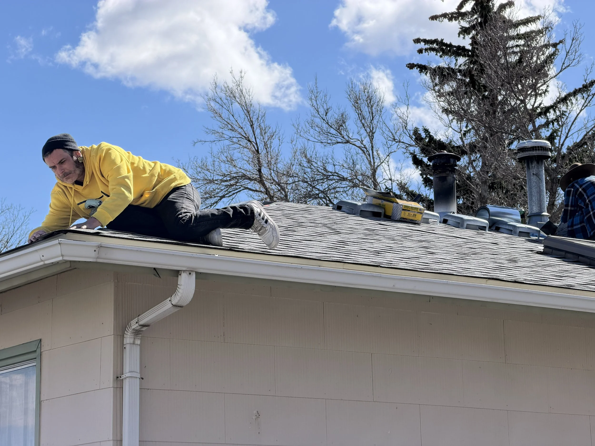 A man and a woman working on the roof of a house during the daytime. The man, wearing a yellow hoodie and a black hat, is positioned on the roof edging, while the woman, in a plaid shirt and hat, is near a chimney vent. There are tools and equipment 