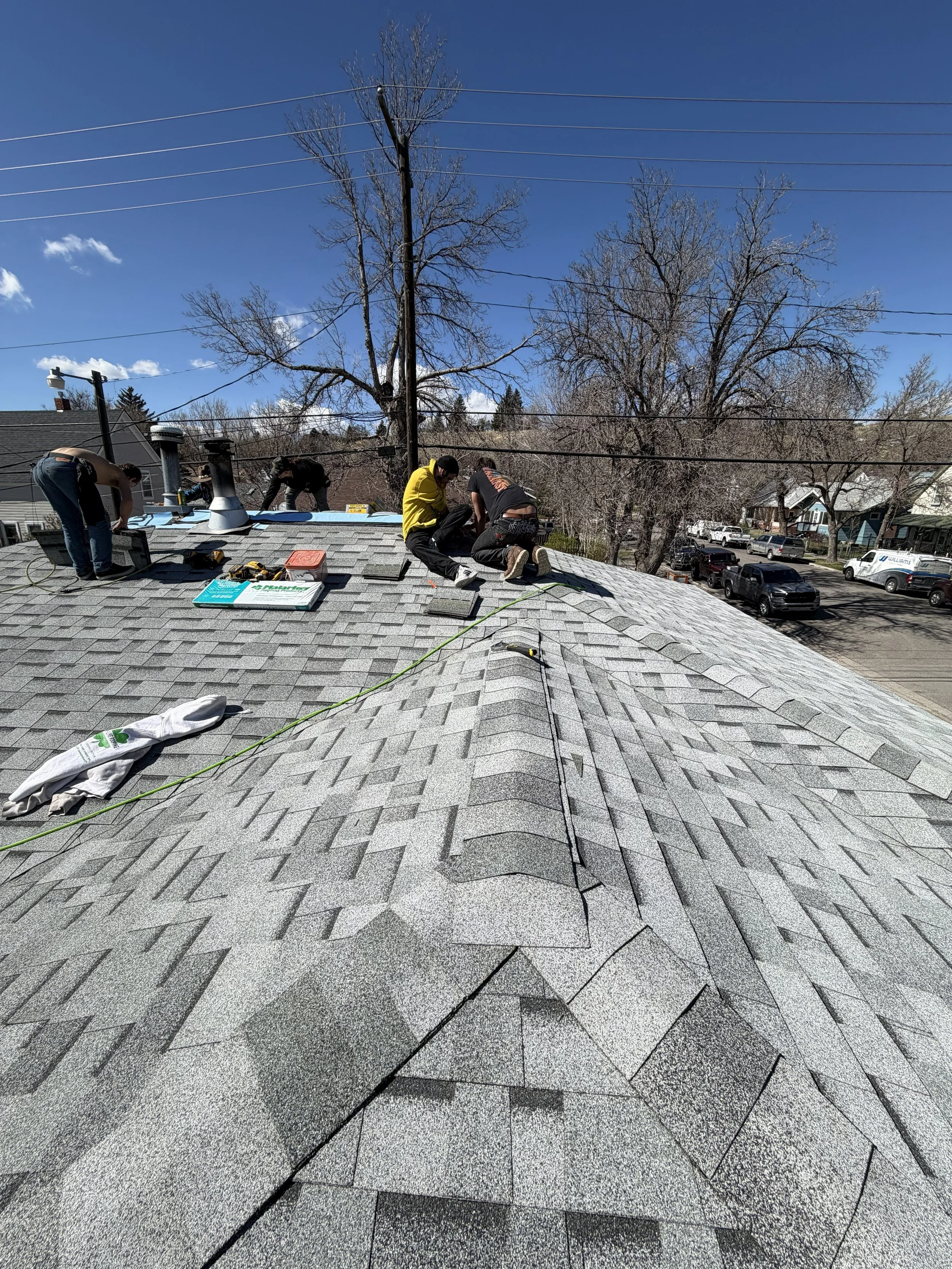 Workers installing new shingles on a residential roof under a clear blue sky with leafless trees and utility lines in the background.